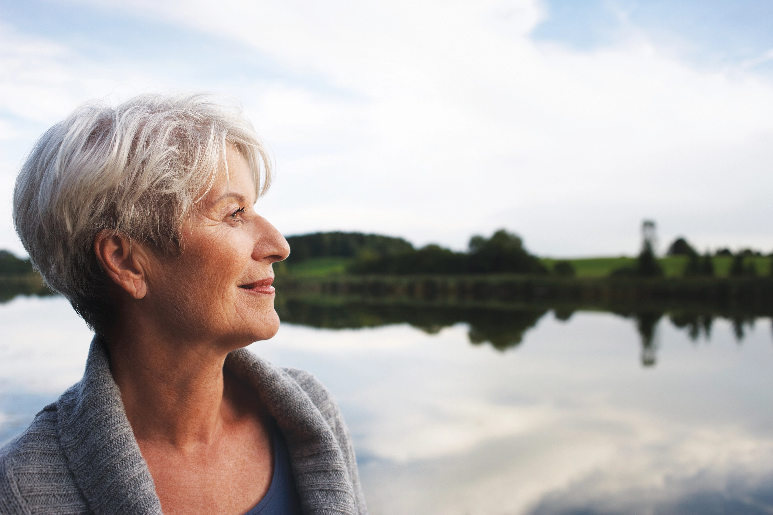 Older woman with short gray hair stands by a calm lake, looking to the side, with trees and a cloudy sky reflected in the water.