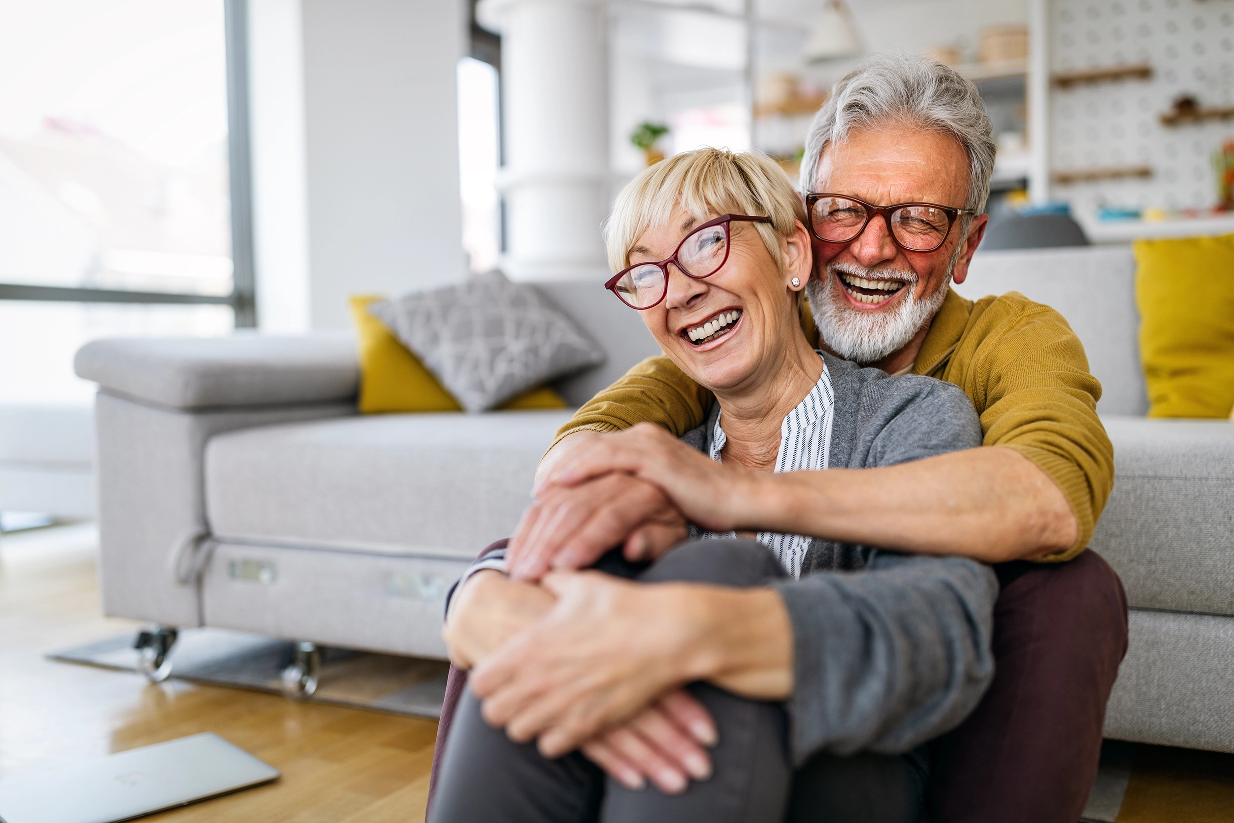 two older people hug each other sitting on the floor of their home